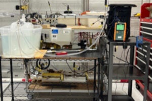 A metallic table holds a translucent water container connected by tubing to a black container on the right. A yellow electronic device measures data. Additional containers and equipment are visible in the background. The setup is part of an experiment on combined offshore floating wind turbines and wave energy converters (WECs).
