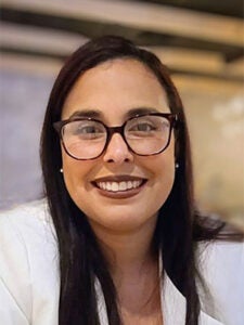 young latino woman with black hair and a white blouse in front of a cafe background smiling with black glasses on
