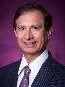 Man smiling in a dark purple suit, white shirt, and purple tie in front of a purple background