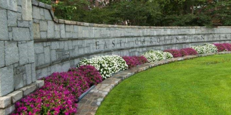 East Carolina university grey brick wall behind flowered shrubbery and a green lawn with trees in the background