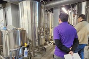 : Two men touring a boiler room. One is in a purple shirt and grey pants with black hair. The other man is in a khaki jacket and denim pants with black hair. 