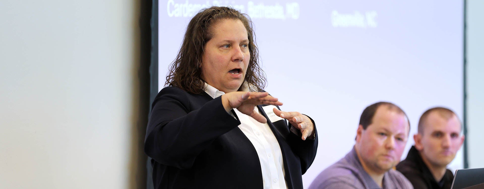 A woman stands, gesturing with her hands as she talks, while two men sit at right and listen.
