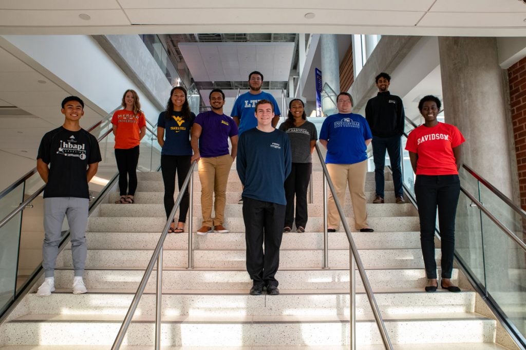 Group of students on the steps inside the Main Campus Student Center wearing various college t-shirts