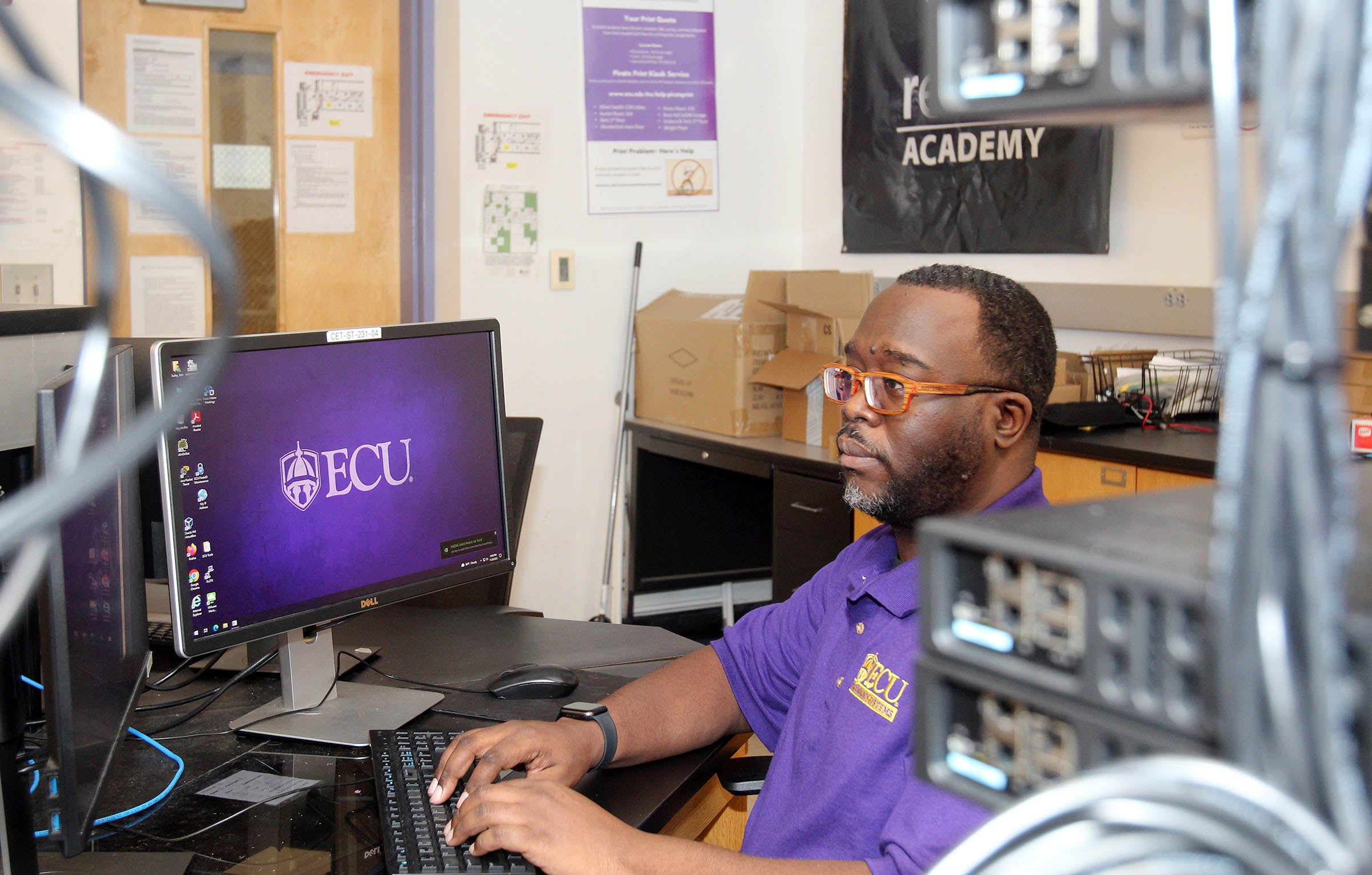A man in a purple shirt sits at a table in a classroom typing on a keyboard while surrounded by computer cables and servers.
