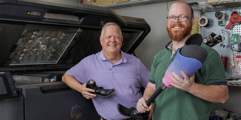 Two men, one at left in a purple shirt and the other in a green shirt, stand next to a large machine that prints three-dimensional objects while holding prosthetic foot attachments.