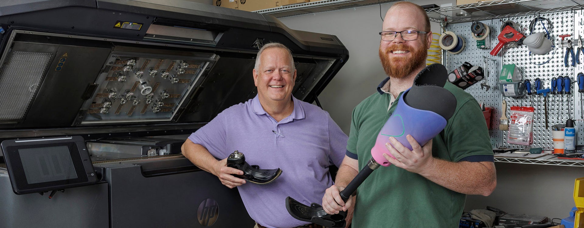 Two men, one at left in a purple shirt and the other in a green shirt, stand next to a large machine that prints three-dimensional objects while holding prosthetic foot attachments.
