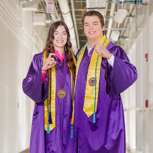 A young women in a purple graduation gown at left, holding her hand and fingers up to form a hook, stands next to a young man wearing a purple graduation gown holding his hand and fingers up to form a hook.