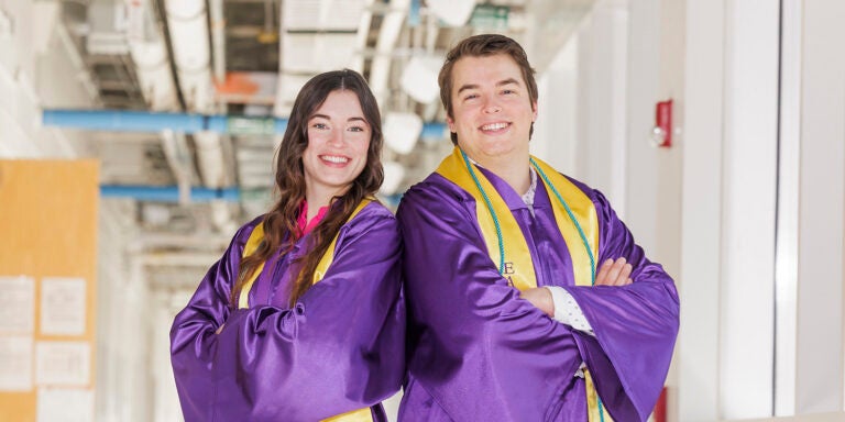 A young woman wearing a purple graduation gown stands at left with her arms crossed next to a young man in a purple graduation gown standing with his arms crossed.