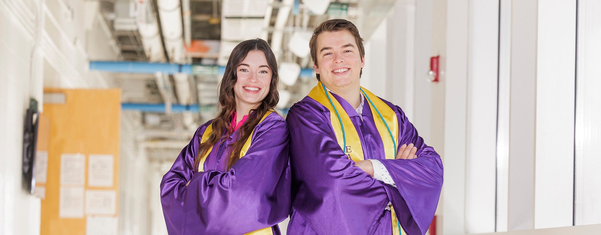 A young woman wearing a purple graduation gown stands at left with her arms crossed next to a young man in a purple graduation gown standing with his arms crossed.