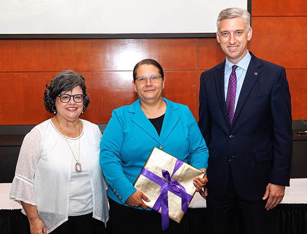 A woman in a blue jacket at center holds a square, wrapped package as she stands between a woman in a white sweater at left and a man in a suit at right.