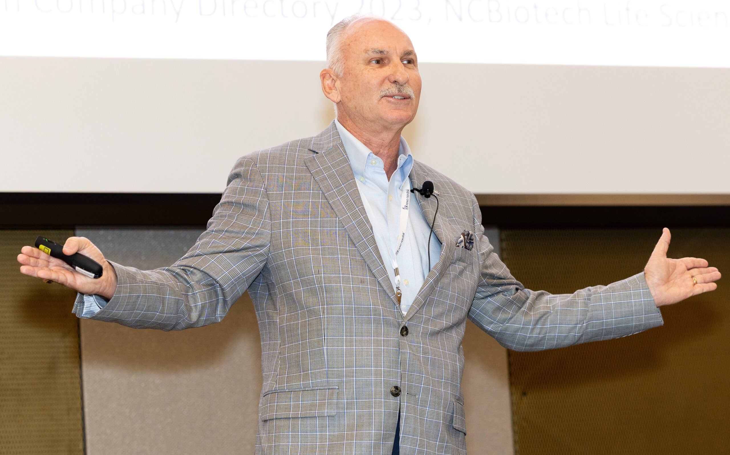 A man in a gray sport coat outstretches both of his arms as he speaks in front of a screen from a stage.