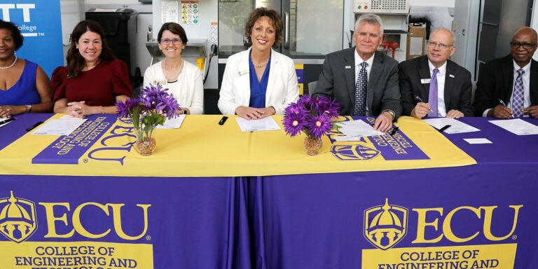 Four women at left sit next to three men at right at a table with a purple tablecloth as they look up while signing papers in front of them.