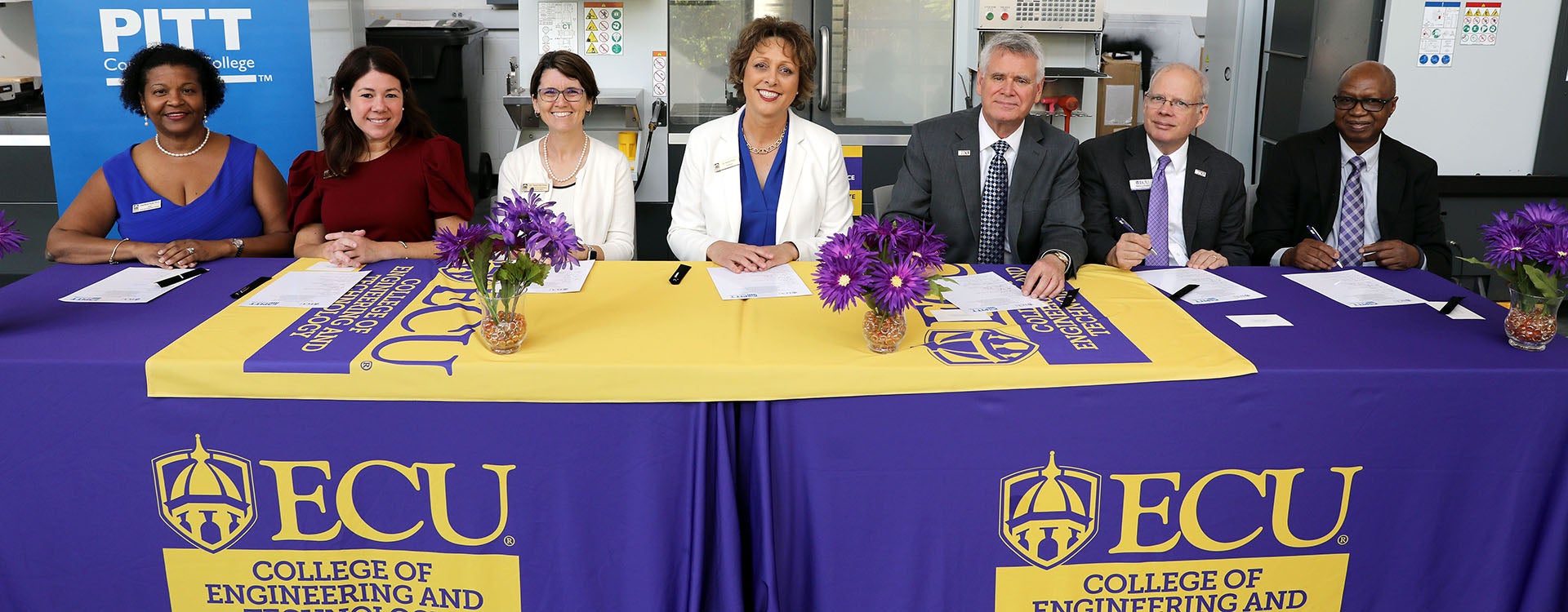Four women at left sit next to three men at right at a table with a purple tablecloth as they look up while signing papers in front of them.