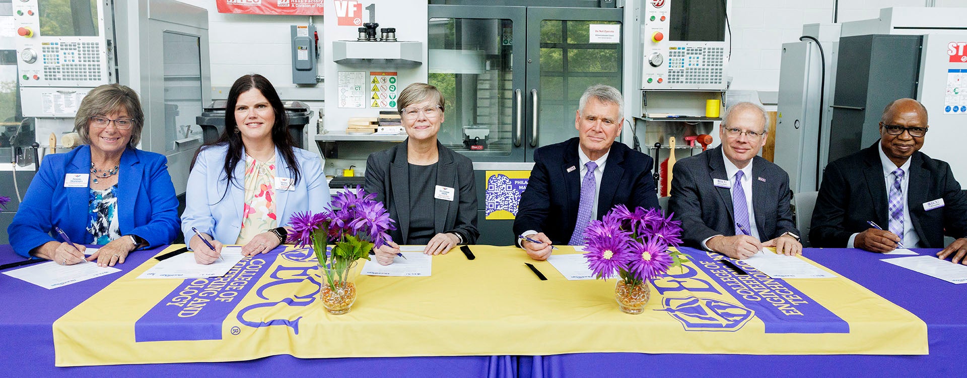 Three women at left sit next to three men at right at a table covered in a purple tablecloth as they sign documents.
