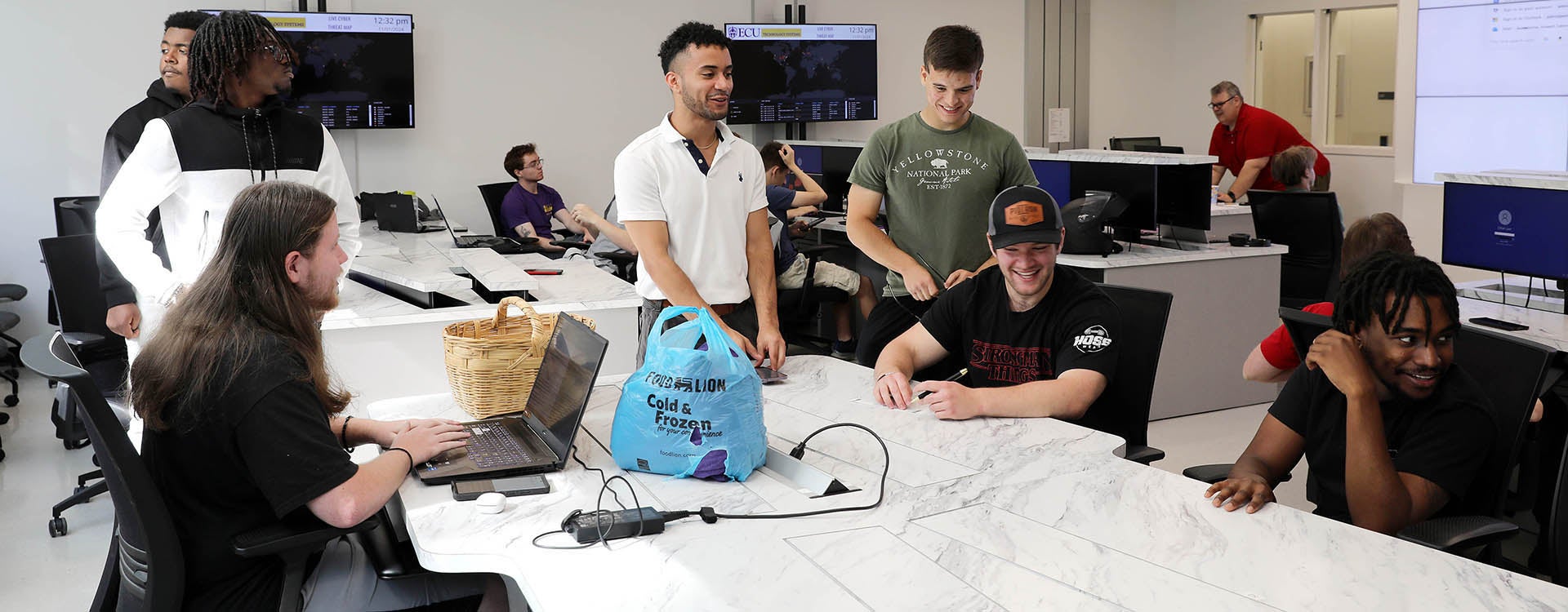 A group of seven students, some sitting at a long desk and some standing around it, talk and smile while one types on a laptop computer.