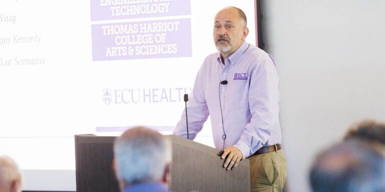 A man in a purple and white shirt stands at a podium and speaks to people who are sitting in front of him.