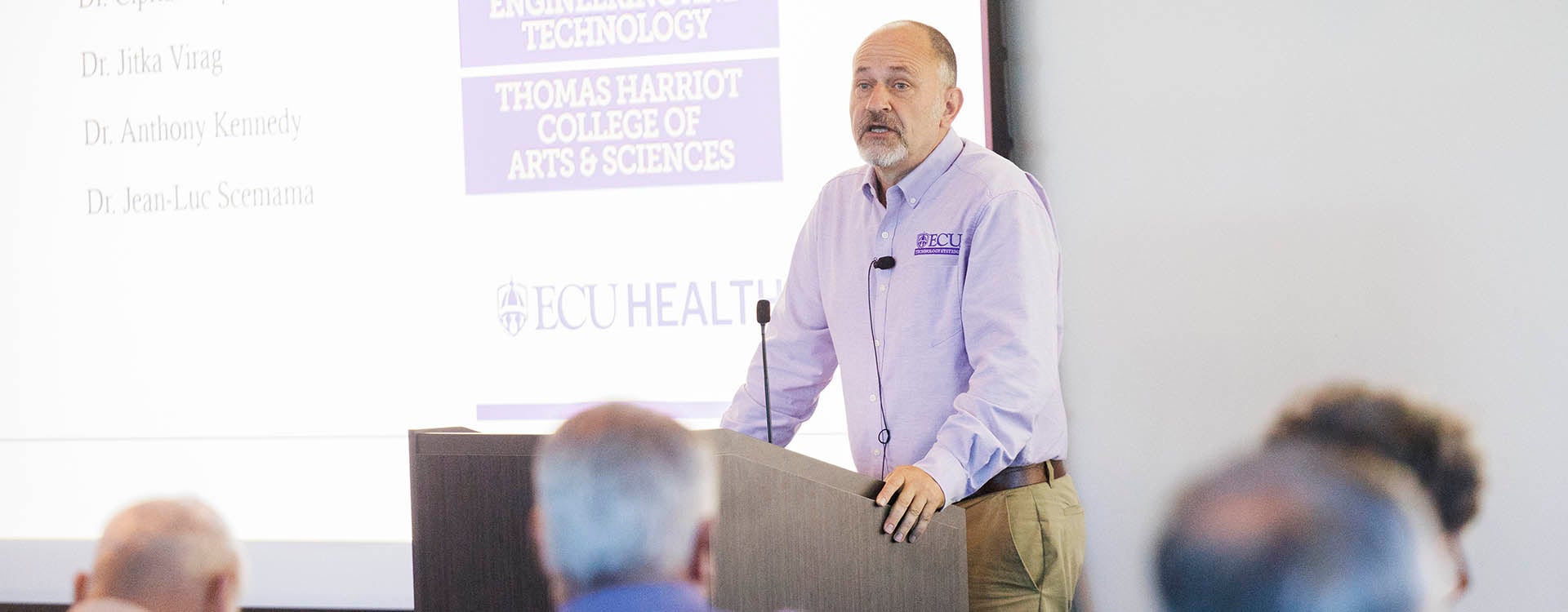 A man in a purple and white shirt stands at a podium and speaks to people who are sitting in front of him.