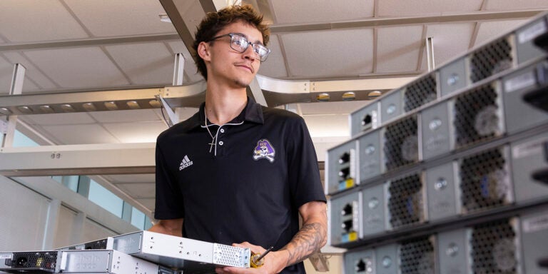 A male student in a black shirt with an East Carolina University logo pulls a computer server from a stack to his right as other servers are stacked to his left.