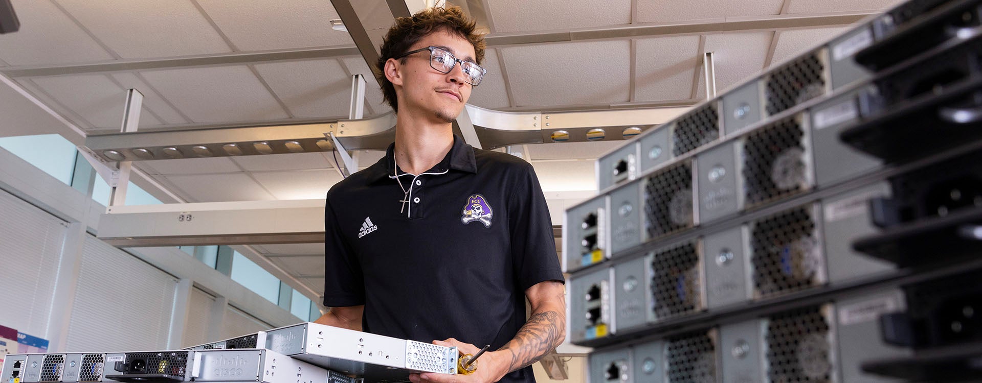 A male student in a black shirt with an East Carolina University logo pulls a computer server from a stack to his right as other servers are stacked to his left.