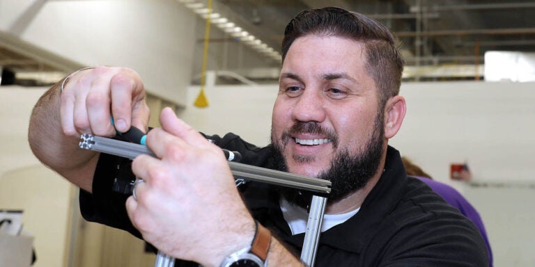 A smiling man in a black shirt uses a small wrench to fasten two pieces of metal.