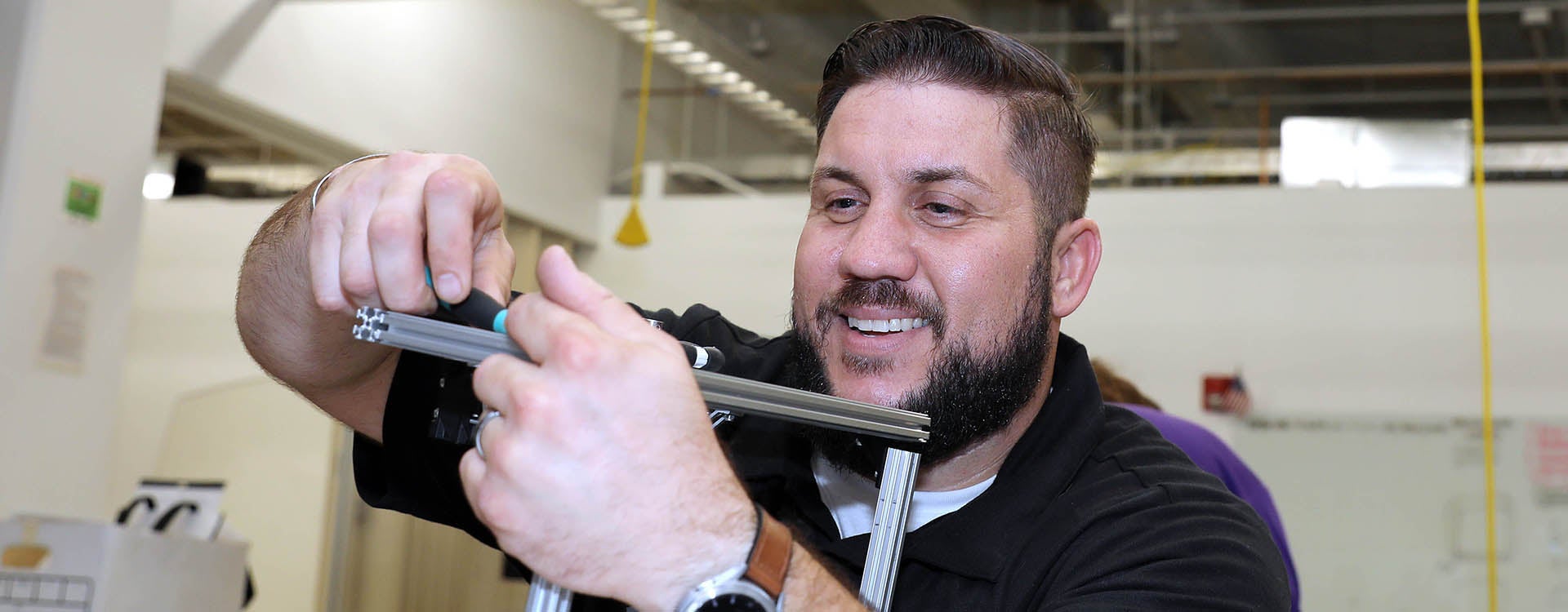 A smiling man in a black shirt uses a small wrench to fasten two pieces of metal.