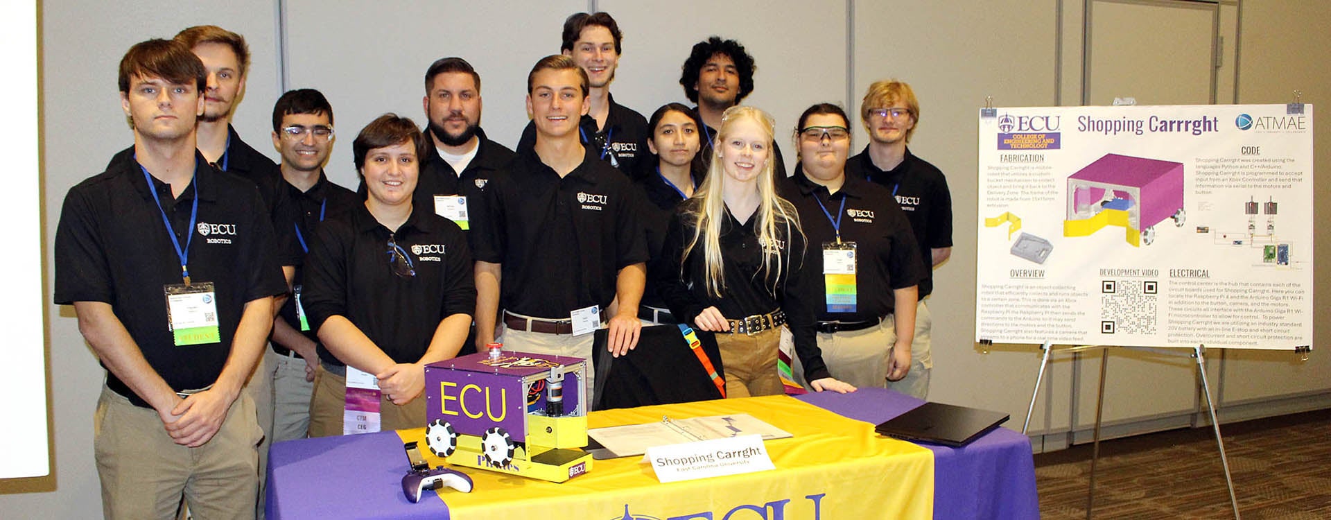 Twelve people in black shirts stand behind a table that includes a purple and gold robot that looks like a small, covered shopping cart. Next to them is a poster that describes how they built the robot. The table is covered with a purple and gold tablecloth.