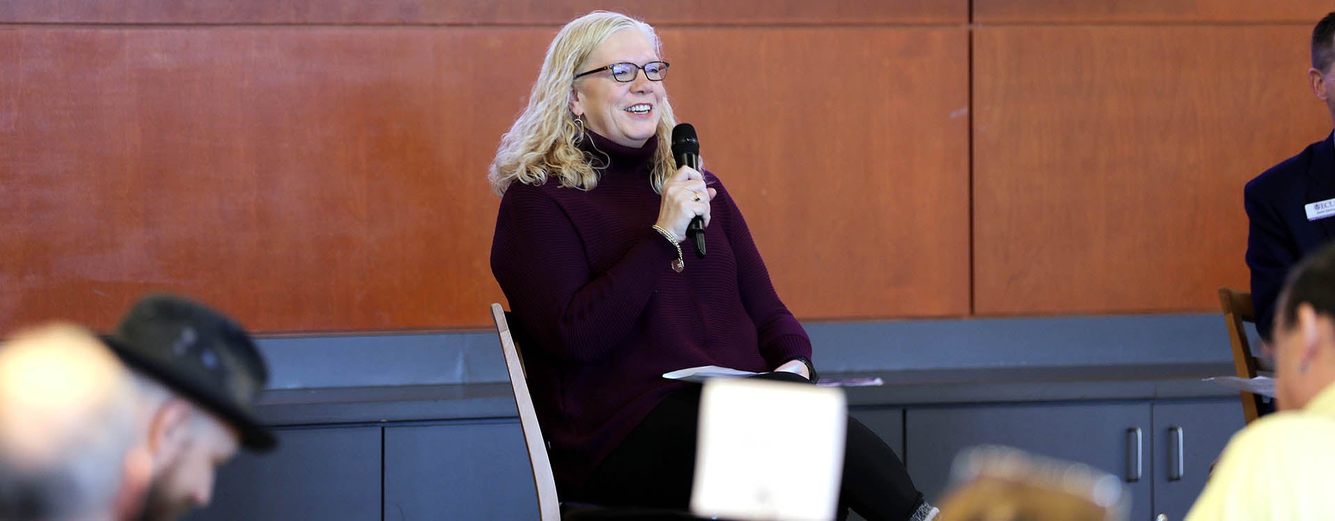 A woman in a sweater smiles as she holds a microphone and speaks as she sits on a chair in front of a group of people.