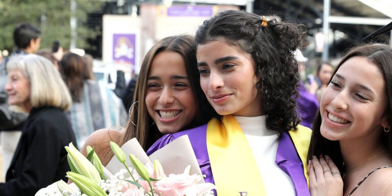 Three young women, the one in the middle with a purple graduation gown holding flowers, smile and embrace as they pose for a photograph.