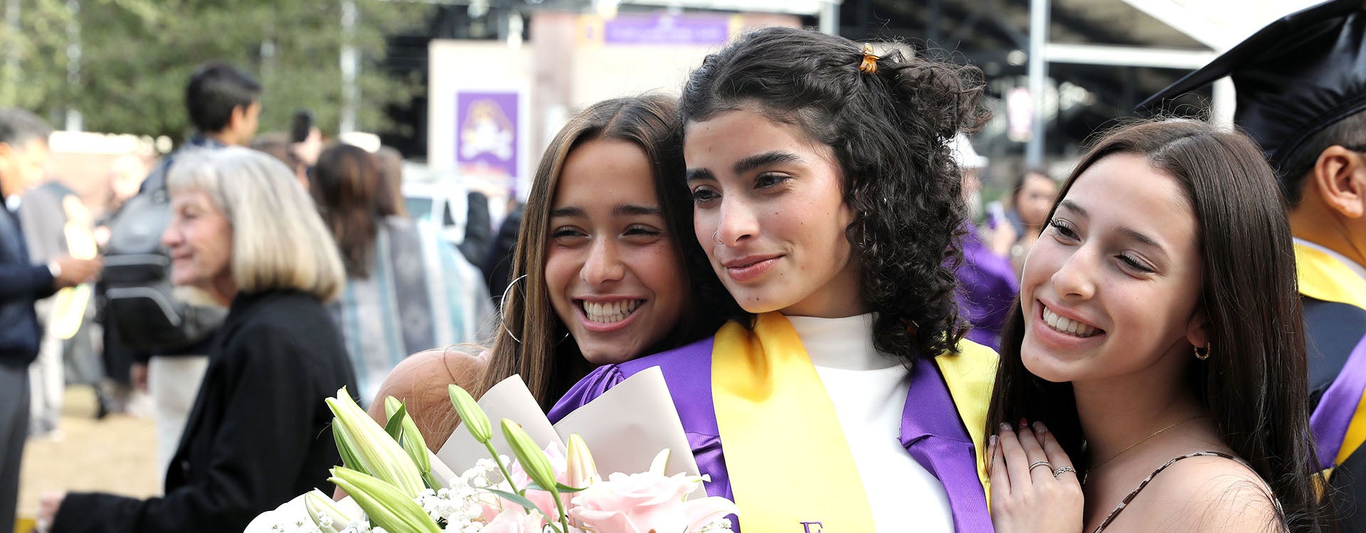 Three young women, the one in the middle with a purple graduation gown holding flowers, smile and embrace as they pose for a photograph.