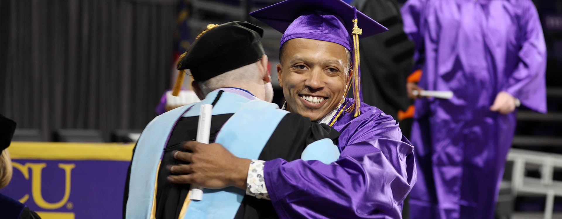 A young man at right in a graduation cap and gown gets a hug from another man in a commencement robe.