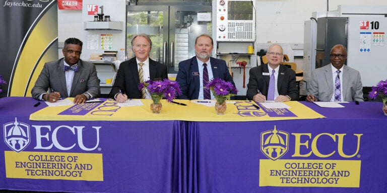 Five men in suits and ties sit at two tables covered in purple with East Carolina University logos on the front.