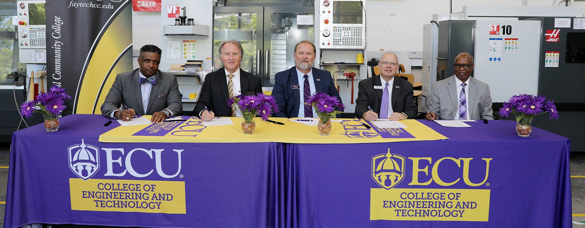 Five men in suits and ties sit at two tables covered in purple with East Carolina University logos on the front.