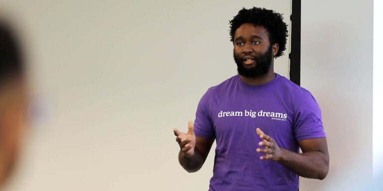 A bearded man in a purple shirt gestures with his hands as he stands at the front of a room and talks.