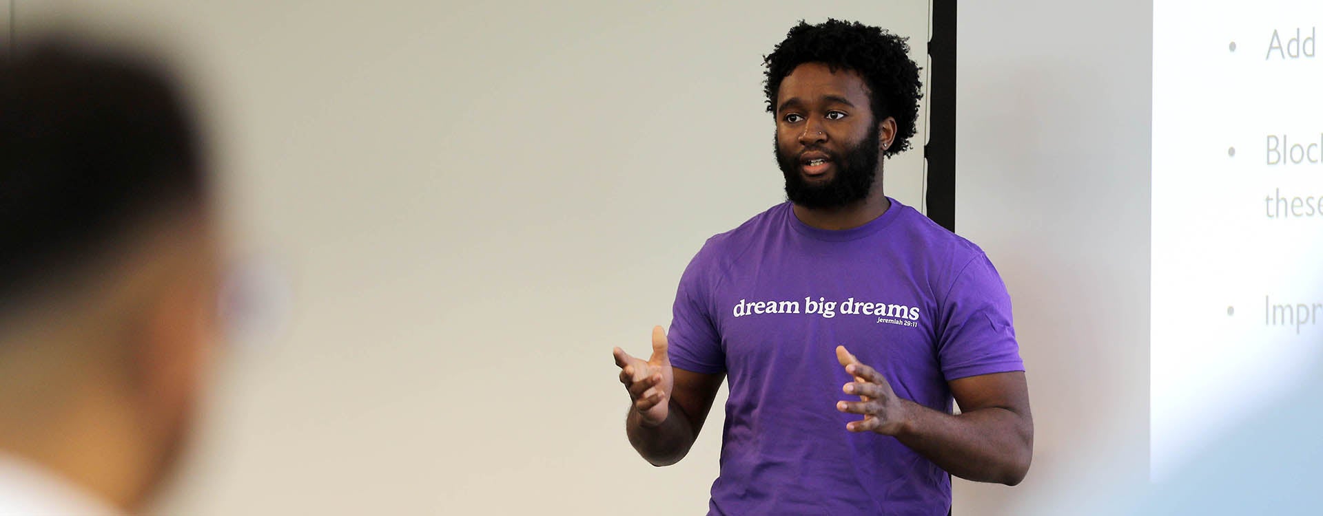 A bearded man in a purple shirt gestures with his hands as he stands at the front of a room and talks.