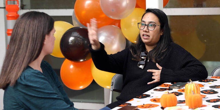 A woman wearing a black shirt sitting at a table gestures with her hand in the air as she talks to another woman sitting at the table with orange, black, yellow and white balloons in the background.