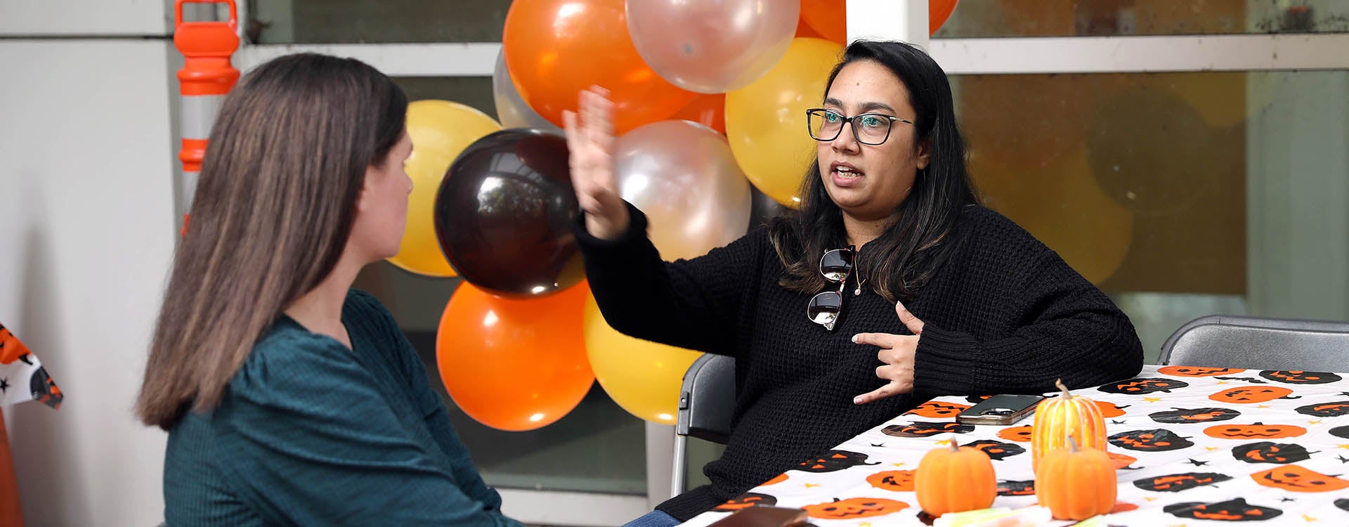 A woman wearing a black shirt sitting at a table gestures with her hand in the air as she talks to another woman sitting at the table with orange, black, yellow and white balloons in the background.