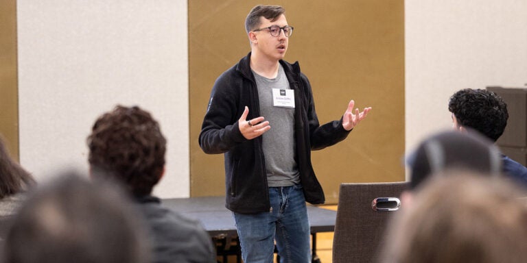 A man wearing a black jacket and gray shirt gestures with his hands as he stands and talks to numerous people seated in chairs to the left and right of him.