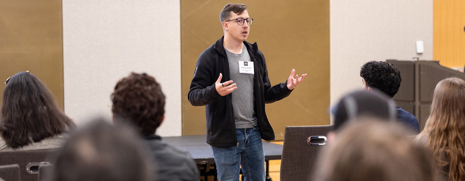 A man wearing a black jacket and gray shirt gestures with his hands as he stands and talks to numerous people seated in chairs to the left and right of him.