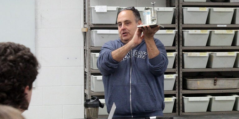 A man in a gray sweatshirt holds up a cylinder to show students sitting at a lab table.