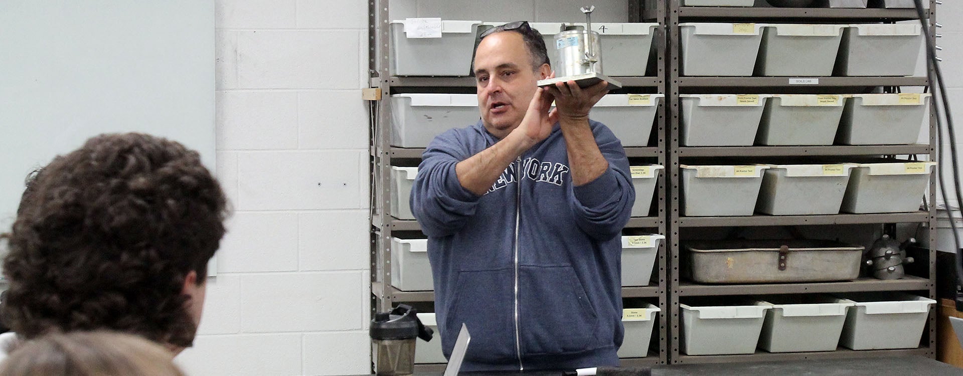 A man in a gray sweatshirt holds up a cylinder to show students sitting at a lab table.