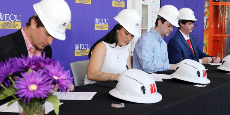 Four people wearing ECU construction hard hats sit at a table and sign documents.