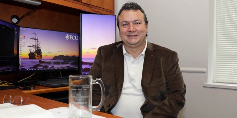 A man wearing a brown sport coat and white shirt sits at a desk in an office in front of two computer screens with an empty glass mug on the desk in front of him.