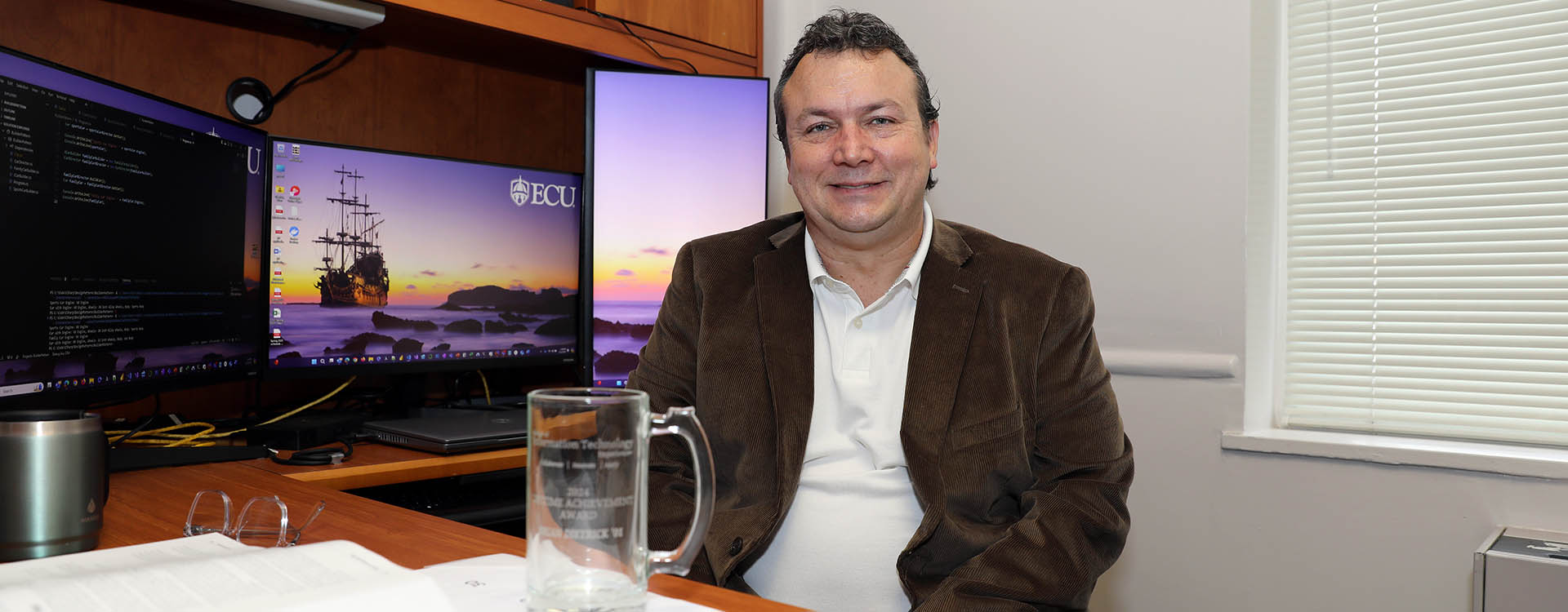 A man wearing a brown sport coat and white shirt sits at a desk in an office in front of two computer screens with an empty glass mug on the desk in front of him.