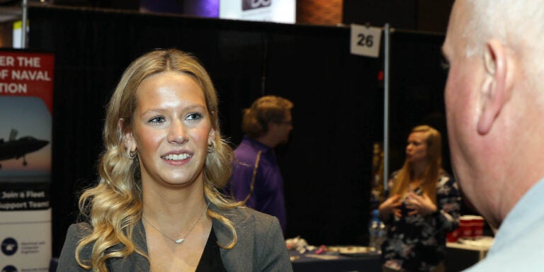 A woman in a gray jacket smiles as she talks with a person at right.