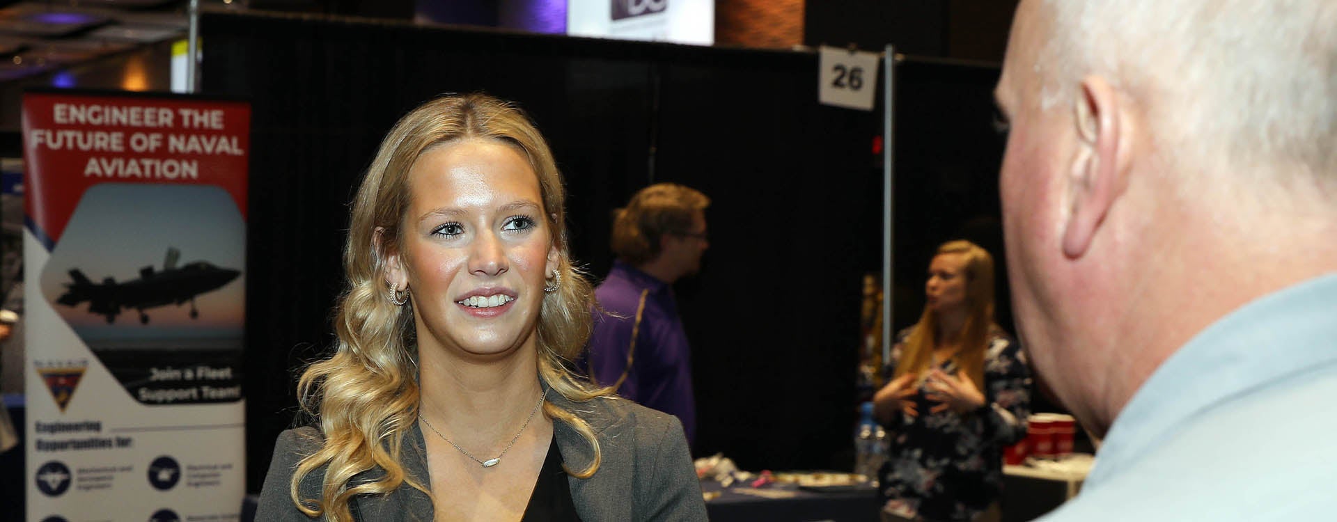 A woman in a gray jacket smiles as she talks with a person at right.