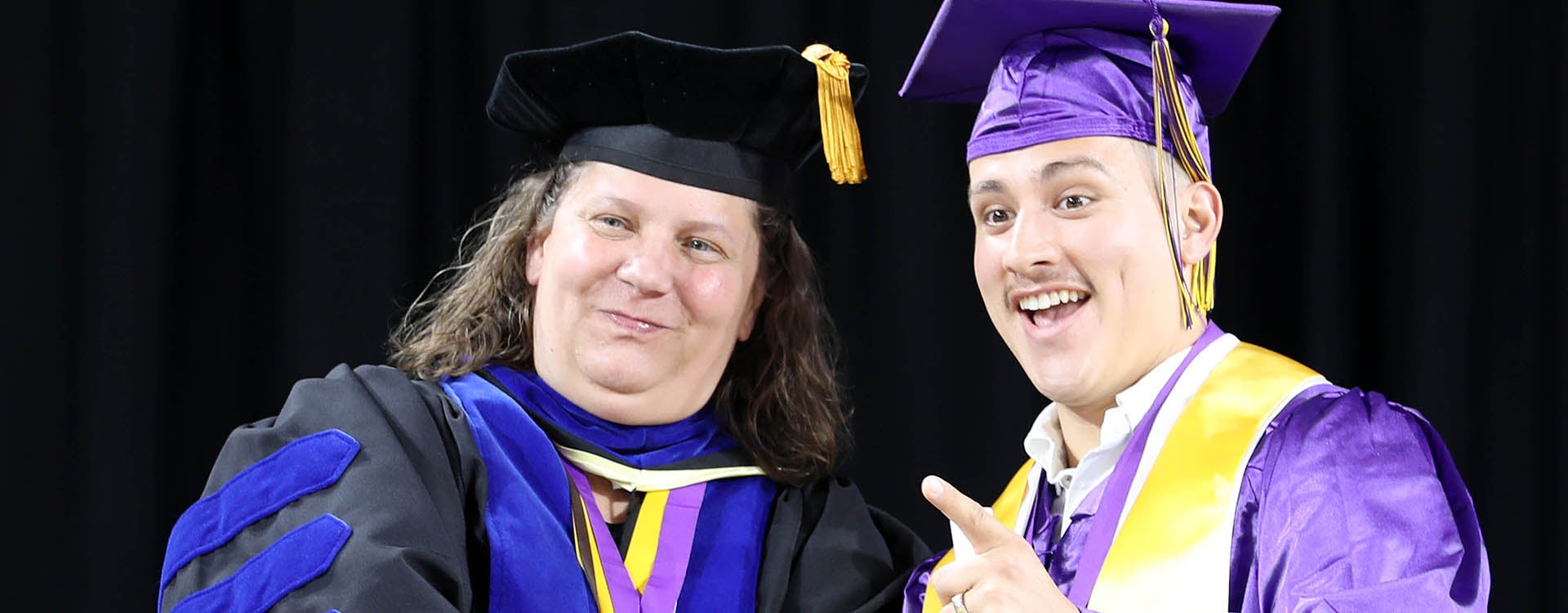 A student at right in a purple graduation cap and gown smiles and points as he stands next to a woman in a graduation robe.