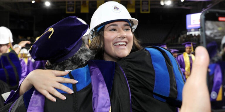 A young woman wearing a white hard hat with an East Carolina University logo smiles as she receives a hug from a woman in a graduation robe.
