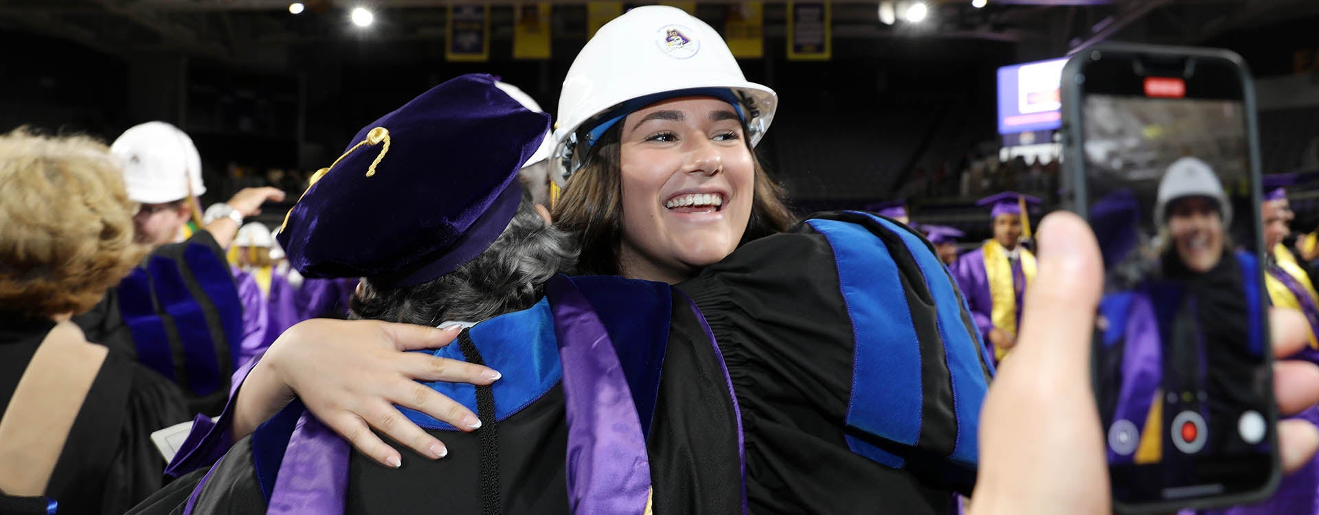 A young woman wearing a white hard hat with an East Carolina University logo smiles as she receives a hug from a woman in a graduation robe.