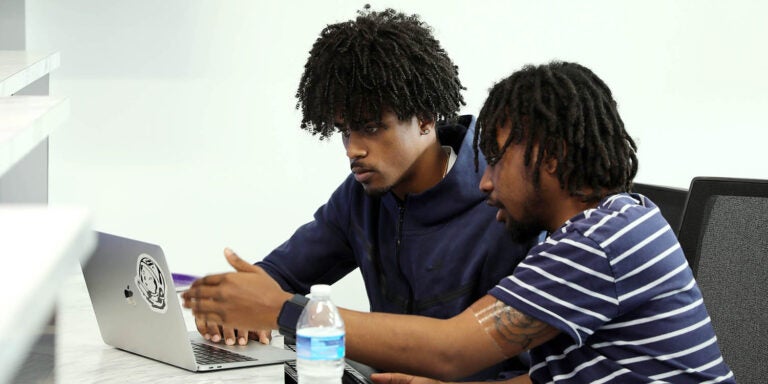 Two young men, one in a short-sleeved shirt and the other in a hooded sweatshirt, sit at a table and look at a laptop computer.