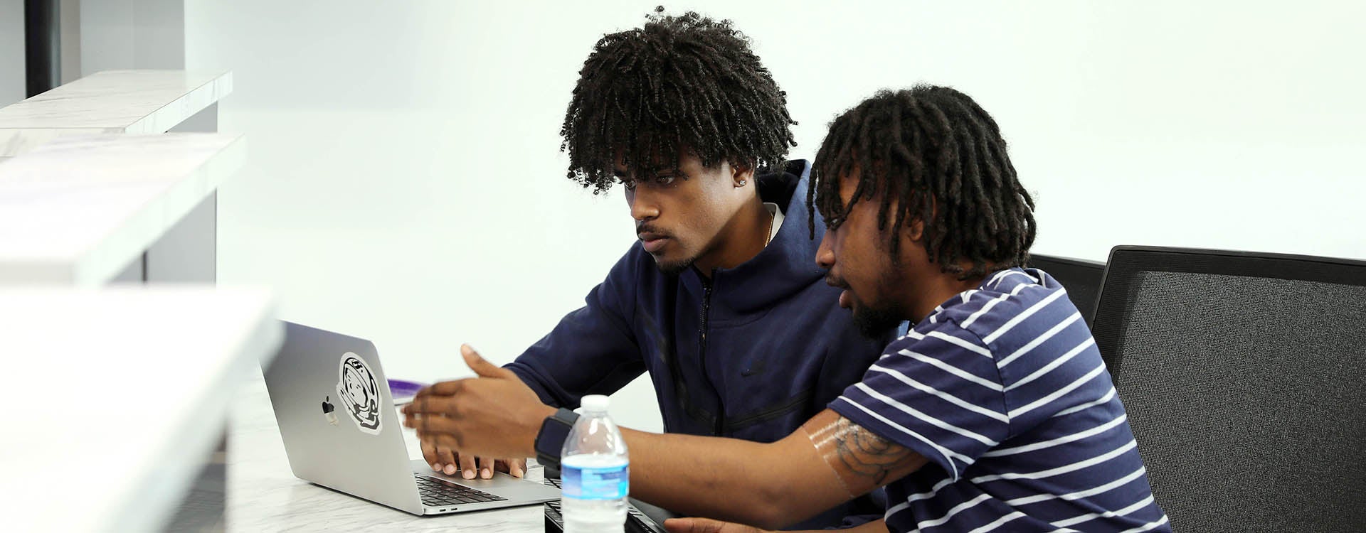 Two young men, one in a short-sleeved shirt and the other in a hooded sweatshirt, sit at a table and look at a laptop computer.